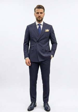 Hombre de pie mirando hacia adelante vestido con un traje cruzado a rayas azul marino, corbata de lunares, camisa blanca y pañuelo de bolsillo.