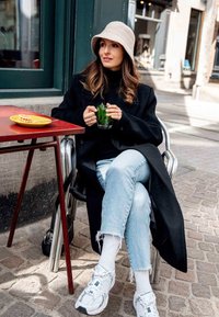 A black coat over a black top, light blue jeans, and white sneakers. A beige bucket hat, holding a glass with greenery, seated at a red table.