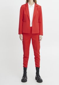 Blazer rouge ajusté et pantalon fuselé assorti en tissu lisse, associé à une chemise blanche boutonnée et à des bottines noires épaisses à lacets.