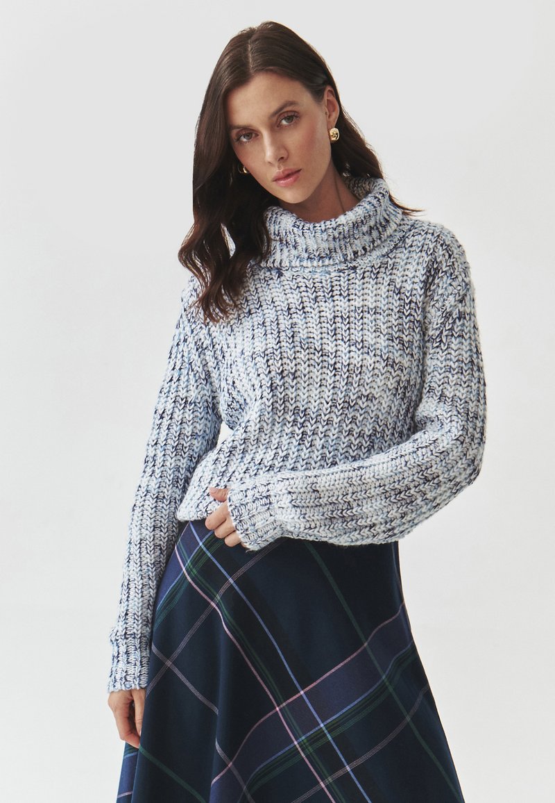 Gestrickter Rollkragenpullover in Hellblau und Weiß mit einer groben Textur. Kombiniert mit einem marineblauen Karorock, der grüne und violette Streifen aufweist.