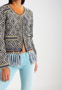 Veste tissée noire et blanche à motifs, avec des accents jaunes et bleus, bordure à franges, deux poches et un col rond. Associée à un jean bleu clair.