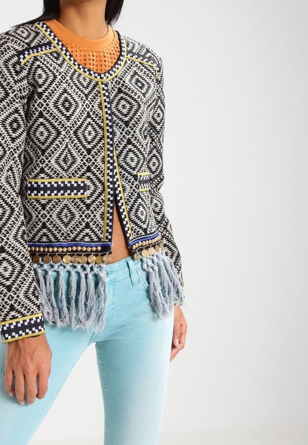 Veste tissée noire et blanche à motifs, avec des accents jaunes et bleus, bordure à franges, deux poches et un col rond. Associée à un jean bleu clair.