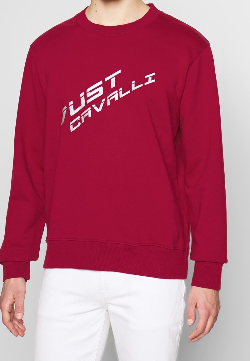 Rode sweatshirt met een ronde hals, lange mouwen en een zilveren grafisch logo "JUST CAVALLI" dat diagonaal op de voorkant is gedrukt.
