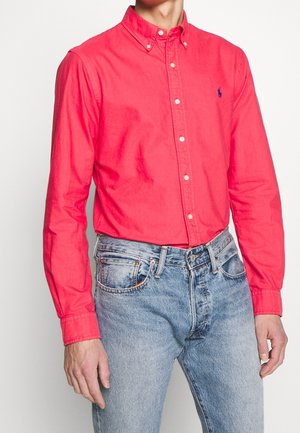 Hombre con una camisa de botones roja brillante metida en unos vaqueros de mezclilla azul claro, sobre un fondo blanco liso.