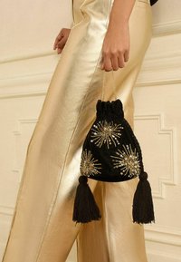 Čierna zamatová taška na stiahnutie so zlatými ozdobami, zdobená strapcami a zaobleným tvarom. Na pozadí kovové zlaté nohavice.