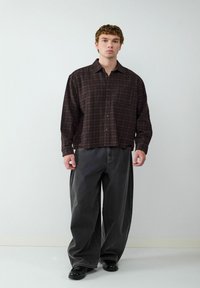 Braun kariertes Hemd mit Knöpfen und einer Brusttasche, kombiniert mit locker sitzenden, dunkelgrauen Hosen und schwarzen Schuhen, vor einem neutralen Hintergrund.