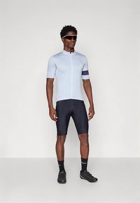 Mann står iført en lyseblå sykkeltrøye med marineblå armring, svarte sykkelsort, svarte sokker, svarte sko og mørke sportsbriller.