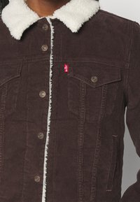 Veste en velours côtelé marron avec col sherpa blanc, fermeture à boutons, poches poitrine à rabat et petite étiquette rouge Levi's sur la poche gauche.