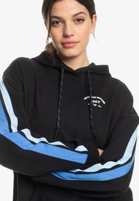Sudadera negra con acentos en rayas diagonales de azul claro y azul marino en las mangas. Cuenta con un logo en la parte frontal y capucha con cordones. Material suave.