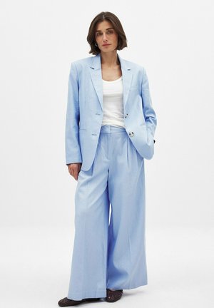 Lyseblå jakkesæt med en skræddersyet blazer og vide bukser, lavet af et glat stof. Båret over en hvid top med store knapper.