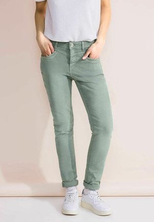 Personne portant un pantalon ajusté vert clair retroussé aux chevilles, des baskets blanches et un haut blanc à manches courtes, debout les mains dans les poches.