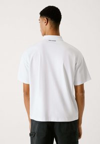 Polo blanco con cuello, mangas cortas y texto de logo sutil en la parte posterior. Tejido suave y corte relajado con dobladillo recto.
