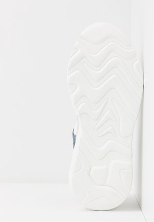 White sneaker sole with zigzag and wavy tread pattern against a white wall and floor background.