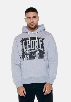 Uomo che indossa una felpa con cappuccio grigio chiaro con la scritta "LEONE 1947" e stampa vintage da boxe, in piedi su uno sfondo chiaro e uniforme.