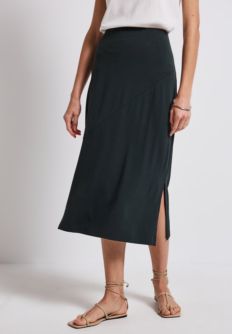 Tmavě zelená midi sukně z měkké látky, s asymetrickým lemem a bočním rozparkem, nošená s béžovými sandály na řemínky.