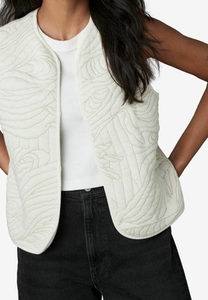 Femme portant un gilet matelassé blanc sans manches avec des coutures abstraites sur une chemise blanche et un pantalon noir, les mains posées sur les hanches.