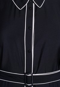 Crna košulja haljina sa dugmadi, s bijelim rubom oko ovratnika, dugmadi i remena s crnom pravokutnom kopčom.