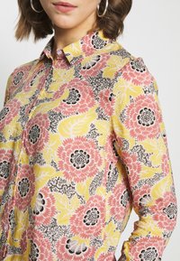 Camisa de botones floral con un fondo amarillo y patrones de flores en rosa y negro, y un cuello. Tela ligera.
