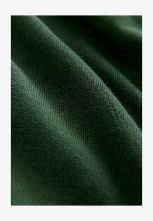 Zeleni pleteni materijal s glatkom teksturom, koji sadrži fine rebra i suptilne varijacije u nijansama. Materijal izgleda meko i fleksibilno.