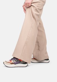 Sport Schuhe, die eine Mischung aus Materialien in Beigetönen, lila und orange aufweisen, mit strukturierten Akzenten und einer dicken weißen Sohle.