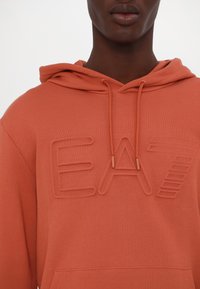 Felpa arancione con un logo testurizzato "EA" sul petto. Presenta un cappuccio con cordino e una tasca a marsupio sul davanti. Tessuto morbido.