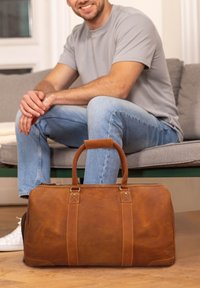 Brun læder duffelbag med to robuste håndtag, syninger som accentuerer, og en lynlås lukning. Glat tekstur og rund form.