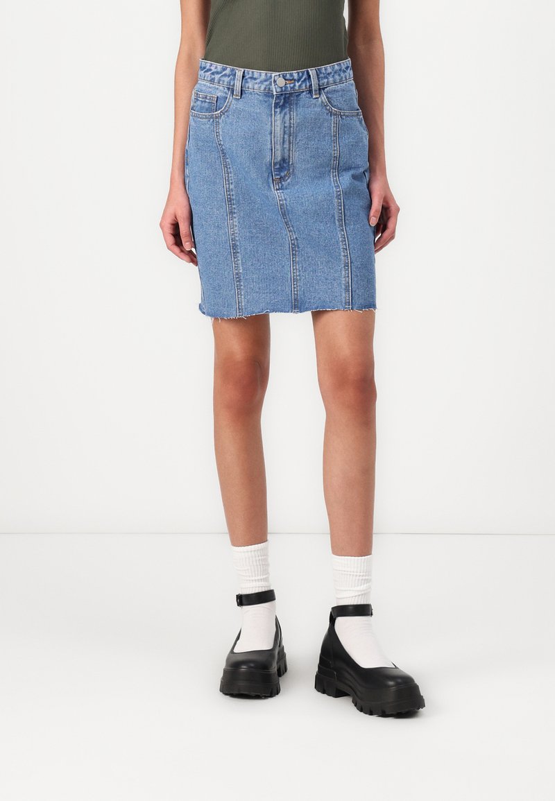 Heledad denimsinine seelik, millel on lihvitud disain, kulunud äär ja eesmistel taskud. Kombineeritud mustade turskete jalatsitega ja valgete sokkidega.