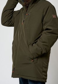 Muž v olivově zelené bundě na zip s kapsou na hrudi, kapsami na ruce a logem na horním rukávu.