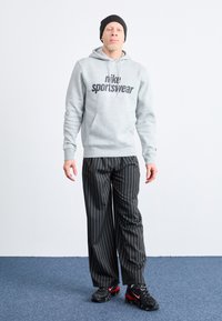 Pilkas džemperis su "Nike Sportswear" logotipu, derinamas su juodomis smulkiai dryžuotomis kelnėmis ir juodais sportiniais batais su raudonais akcentais. Ant galvos uždėta juoda kepurė.