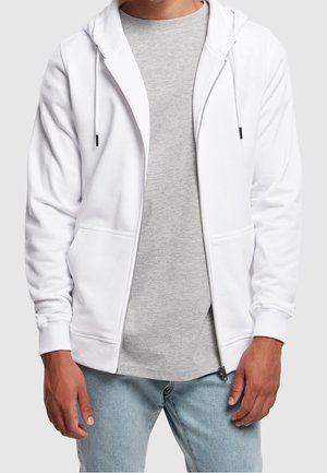 Homme portant un sweat à capuche blanc avec fermeture éclair sur un t-shirt gris et un jean bleu clair, debout devant un fond clair uni.