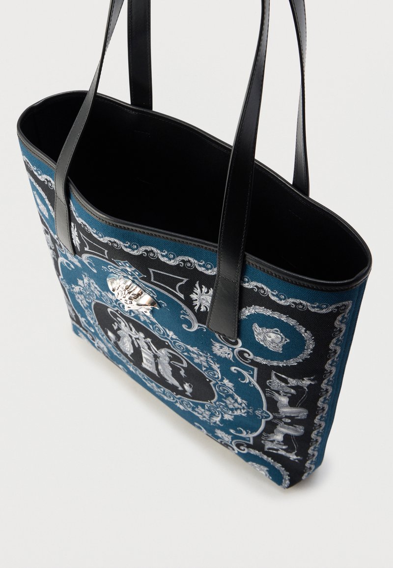Sac fourre-tout en toile bleue avec des détails noirs, présentant des motifs argentés complexes et un accent décoratif en métal argenté à l'avant.