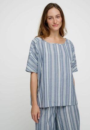 Vrouw met een lichte huid en bruin haar, die een losse blauw-wit verticaal gestreepte kortärmelige blouse en bijpassende broek draagt, staand tegen een witte achtergrond.