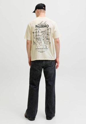 Uomo che indossa una maglietta color crema con schizzo nero della città di Palermo e testo, jeans scuri e cappello nero, di spalle su sfondo semplice.
