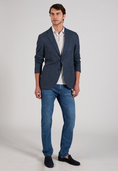 Man stands wearing a dark blue blazer, white shirt, blue jeans, and dark loafers against a plain grey background.