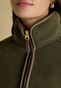 Detailná fotografia osoby v olivovo zelenej fleecovej bunde s hnedými okrajmi a zlatým zipsom, ktorý je čiastočne zipsovaný až po krk, pričom má na sebe zlatú kruhovú náušnicu.