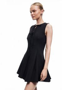 Sort ærmeløs kjole med tætsiddende overdel og flagrende nederdel, der har en bindedetalje ved halsen og et glat, struktureret stof.