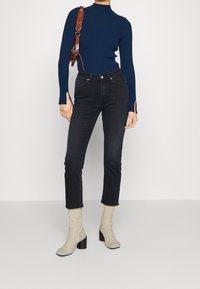 Navy ribbed long-sleeve top, black high-waisted jeans, light-colored knitted socks, and dark block-heeled shoes, with a brown strap handbag.