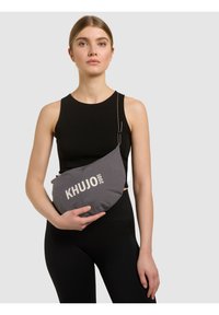 Femme en haut noir sans manches et leggings tenant un sac bandoulière gris avec le logo "KHUJO 2001" sur un fond clair.
