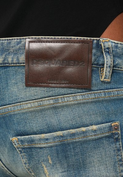 Κοντινό πλάνο μπλε τζιν με καφέ δερμάτινο ταμπελάκι DSQUARED2 πάνω από την πίσω τσέπη και λεπτομέρειες φθαρμένου υφάσματος.