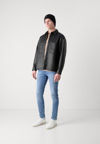 Camisa chaqueta de cuero sintético negra con grandes bolsillos en el pecho, llevada sobre una camiseta beige, combinada con unos pantalones vaqueros azules y zapatillas blancas. Gorro de punto negro.