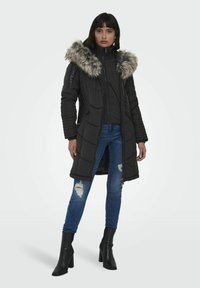 Sort polstret frakke med en faux fur hætte, diamant quiltet mønster, lynlås foran og sidelommer; parret med slidte jeans og hælede støvler.
