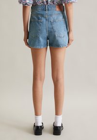 Lichte blauwe denimshorts, hoog in de taille met een relaxed fit, voorzien van twee achterzakken en onafgewerkte zoomranden. gedragen met zwarte schoenen en witte sokken.