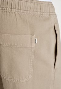Beige bomuldsbukser med elastisk talje, sidelommer og baglommer, samt et lille mærke på baglommen, der viser brandnavnet.