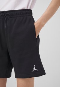 Gros plan d'une personne portant un short noir avec une ceinture élastique et un logo sportif blanc près de l'ourlet, main dans la poche.