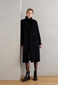Manteau noir en laine à double boutonnage avec de larges revers, poches latérales et coupe mi-cuisse, associé à des bottines noir à lacets.