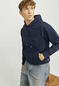 Marineblå hættetrøje i blødt stof med en frontlomme og snøre i hætten. Ordet "ORIGINALS" er embossed på brystet. Denim jeans.