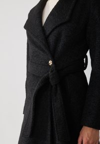 Black wool coat with a textured finish, shawl collar, and a decorative gold button accent. Includes a tied waist belt for adjustable fit.