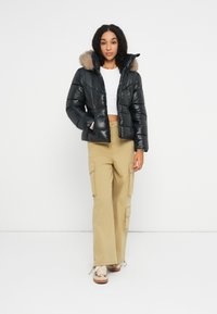 Frau in einer schwarzen Funktionsjacke mit Fellkapuze, weißem Crop-Top, beigen Cargo-Hosen und beigen Sneakers, die vor einem schlichten weißen Hintergrund steht.