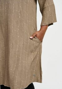 Beige tunikaklänning med en texturerad, skrynklig mönstring, med trekvartsärmar och sidofickor, tillverkad av lätt tyg.