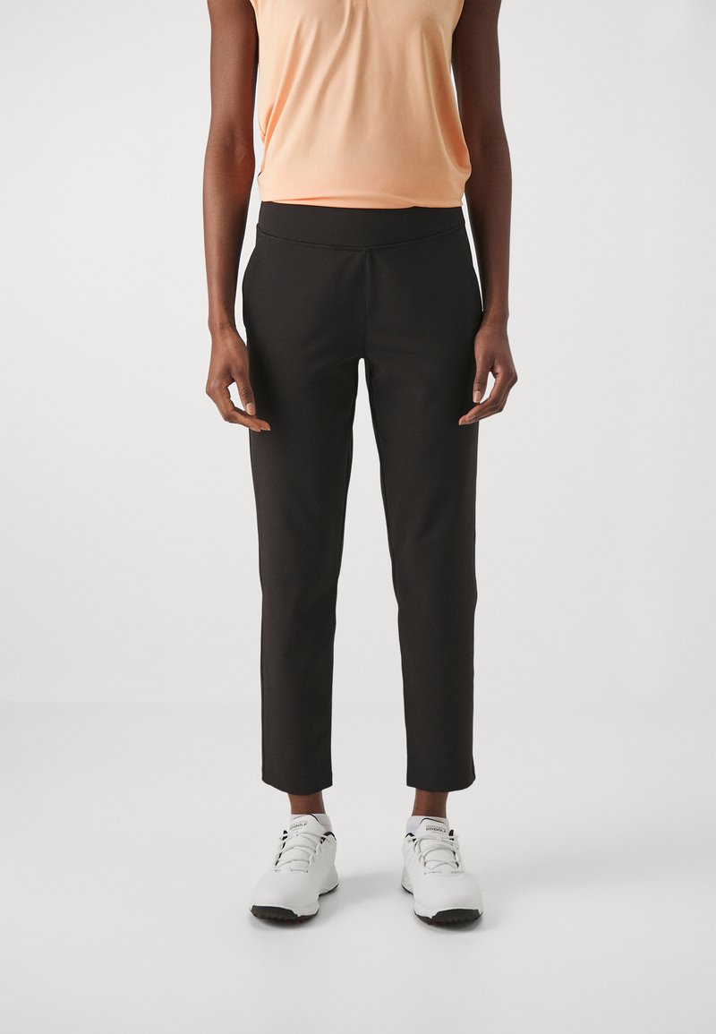 Black tailored pants with a smooth texture, cropped length, and an elastic waistband, paired with white athletic shoes and a peach top.